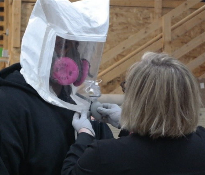A woman spraying into a hood that a man is wearing over his head. The man is also wearing a respirator.